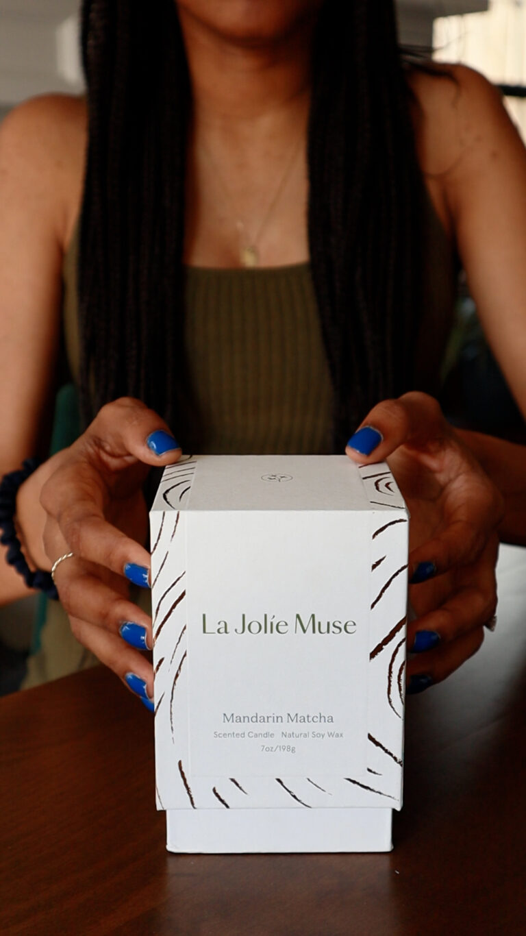 Hands holding a white cardboard box that reads "La Jolie Muse Mandarin Matcha Scented Candle"
