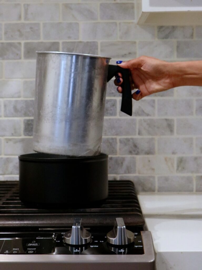 Placing a metal pitcher into a large pot on top of the stove to set up a double boiler for melting candle wax.